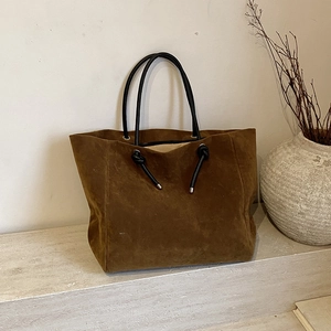 Minimalist dark brown suede tote bag with black circular handles and knot details on a beige stand