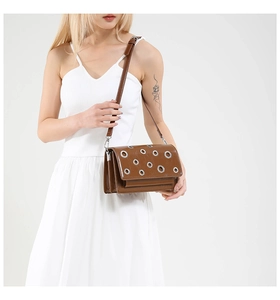 Detail of a brown shoulder bag with silver eyelets worn by a model in a white dress