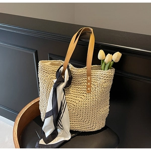 Woven straw tote bag with brown leather handles and decorative silk scarf on a wooden chair