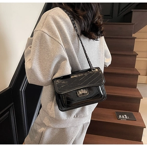 Detail of black glossy quilted chain bag with V-shaped stitching and metal lock on a model
