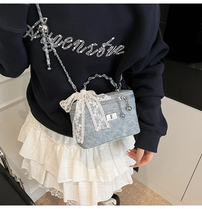 Detail of light blue denim box bag featuring a large white lace bow and silver chain strap