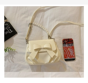 Detail of cream patent leather crossbody bag with bow and pearl chain strap on white fabric