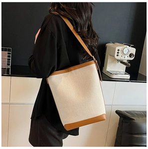Chic beige canvas bucket bag with brown leather trim shown on shoulder in a modern setting