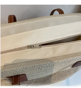 Detail of a woven straw tote bag featuring a smooth metal zipper and brown leather handle
