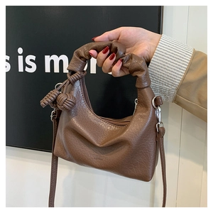 Detail einer braunen halbmondförmigen Falten-Tasche mit Ledertextur und gewebtem Anhänger