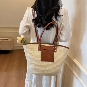 Straw tote bag with brown leather handles and pocket shown on a shoulder in a warm room