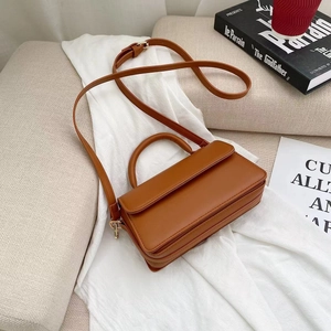 Minimalist caramel brown square crossbody bag with top handle and gold hardware on a sofa