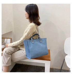 Blue denim tote bag with metal chain strap and top handle shown on a casual seated model