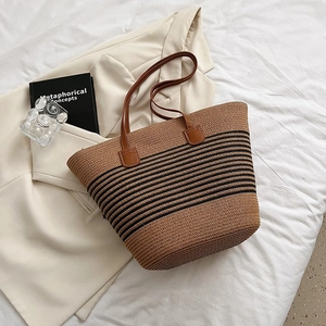 Close-up of woven straw tote bag with black stripes and brown leather handle on soft fabric