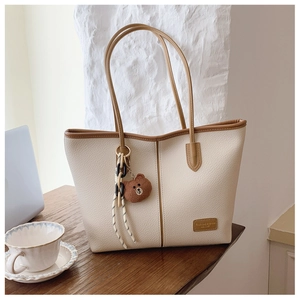 Detail of cream tote bag with litchi grain texture, brown leather tag and plush bear charm