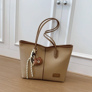 Fashion khaki tote bag with plush bear charm and brown trim on a light wood floor