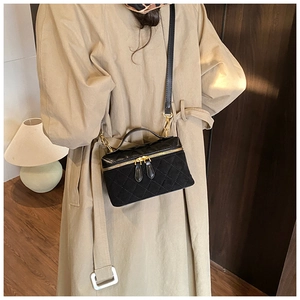 Détail d'un sac vanity noir matelassé avec finitions dorées porté sur un trench-coat kaki