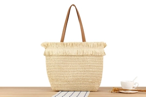 Detail view of woven tote bag with fringe trim and brown handles on a wooden table
