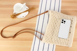 Detail of woven cream crossbody bag with leather strap next to a mobile phone for scale