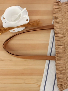 Handmade woven straw tote bag with brown PU straps and fringe detail on wood background