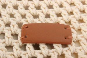 Detail of a brown leather label stitched onto an off-white woven bag with mesh pattern