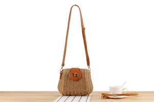 Detail of woven straw crossbody bag with leather flap and metal lock on wooden table