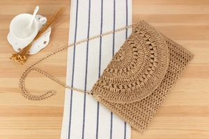 Close-up of a brown crochet straw envelope bag with a gold chain strap on a wooden desk