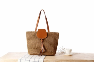 Detail of paper straw woven tote bag featuring brown leather flap and tassel on wooden table