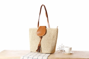 Detail of beige straw woven tote bag featuring brown leather flap and tassel on wood surface