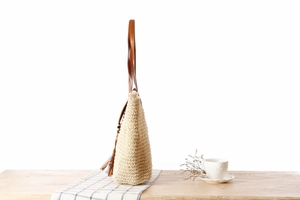 Detail of beige paper rope woven bag with brown leather handle and tassel on wooden table