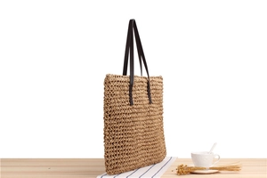 Detail of woven straw tote bag with black handles on a minimalist wooden table