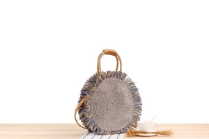 Detail of round straw bag with blue fringes and leather handles on wooden surface