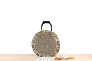 Close-up of circular woven paper straw bag featuring long bicolor fringe and black handles