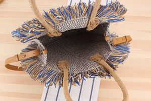 Detail view of woven straw tote bag with long fringe trim and brown leather shoulder strap