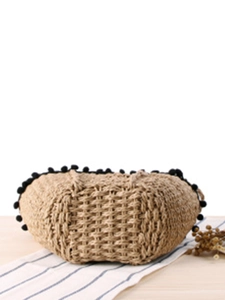 Straw beach tote with black pom pom trim on a wooden table with striped cloth