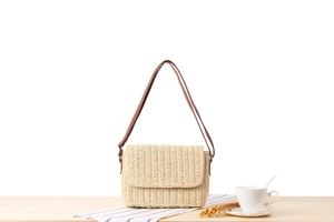 Detail of hand-woven cream straw crossbody bag with brown strap on a rustic wooden table