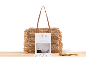 Close-up of woven straw tote bag with side tassels and leather straps on a wooden table