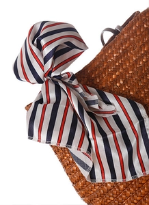 Dettaglio di un foulard di seta a righe rosse e blu annodato al manico di una borsa in paglia