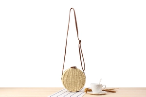 Detail of round straw crossbody bag with brown leather strap and woven texture on wood