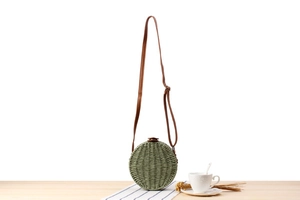 Close-up of green round woven straw bag with leather strap on wooden table surface