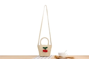 Detail of a beige straw woven bag with decorative red cherry accents on a wooden table