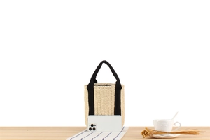 Dettaglio della borsa a secchiello intrecciata beige con manici neri accanto allo smartphone