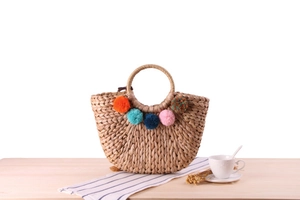 Detail of handmade woven straw bag with colorful pom-poms and round handles on wood table