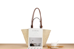 Detail of paper straw tote bag with brown leather handles on a minimalist wood surface
