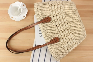 Detail of woven straw tote bag with brown leather handles on a minimalist wooden table