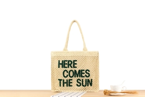Detail of light beige woven tote bag with green Here Comes The Sun embroidery