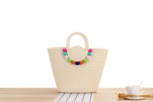 Détail d'un sac cabas en coton tressé crème avec pompons colorés sur une table en bois