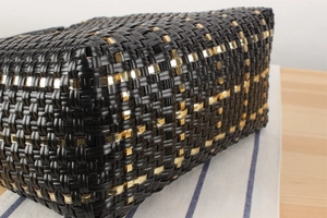 Détail du sac cabas tressé noir avec des accents métalliques dorés sur une surface en bois