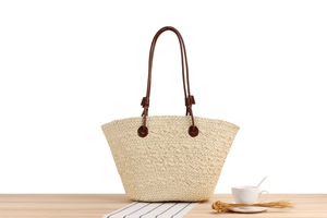 Detail of a beige woven tote bag with slim dark brown handles on a light wooden surface