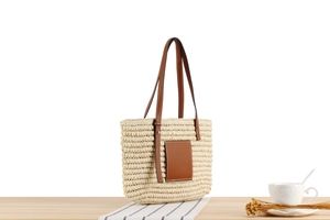Detail of woven straw tote bag with brown leather straps on a light wood table