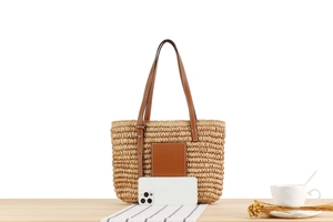 Detail view of woven straw tote bag with brown leather patch and smartphone for scale