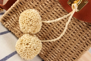 Detail of straw woven bag with leather flap, gold metal lock and cream decorative pom-poms