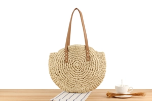 Detail view of a round woven straw beach bag with brown leather handles on a wooden table