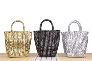 Détail de sacs cabas tissés en doré, argenté et noir posés sur une surface en bois clair