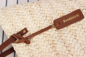 Detail of woven straw bag with dark brown leather strap and golden logo pendant